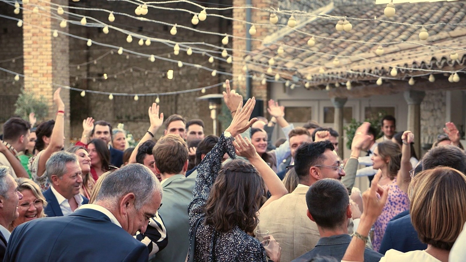 Piste de danse pleine sous des guirlandes lumineuses lors d'un mariage, invités les bras levés pendant le set du DJ