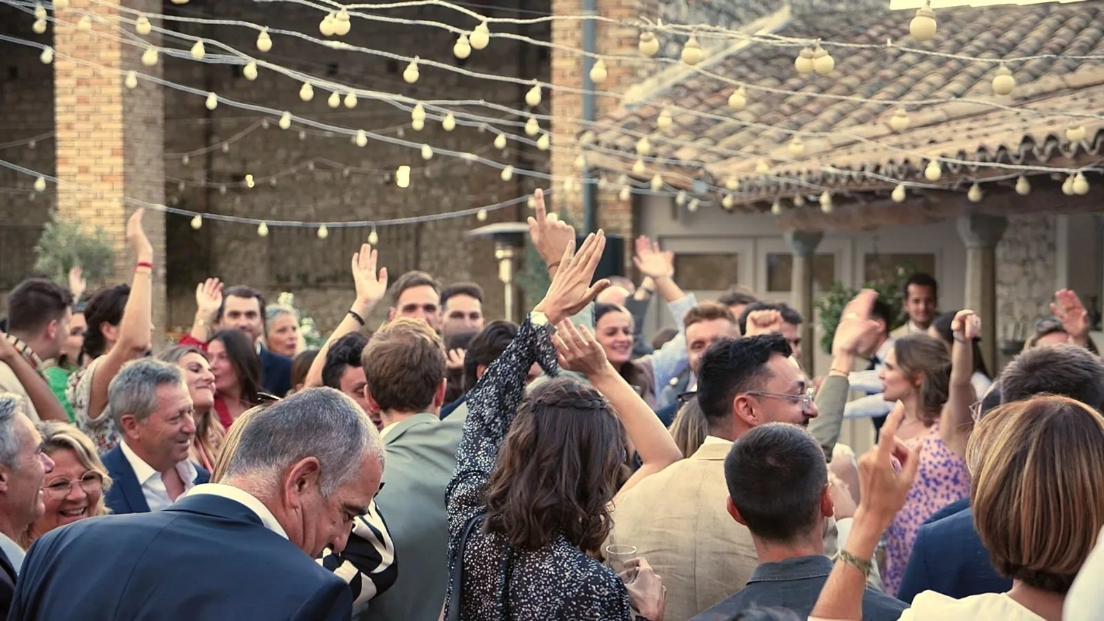 Piste de danse pleine sous des guirlandes lumineuses lors d'un mariage, invités les bras levés pendant le set du DJ